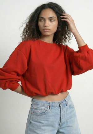 Ung kvinde med krøllet mørkt hår iført en rød cropped sweatshirt og lyseblå jeans, rører ved håret og kigger til siden.
