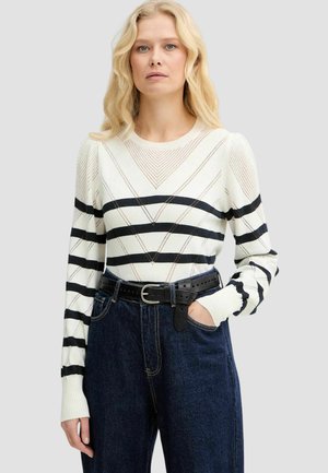 Donna bionda che indossa un maglione bianco a righe nere e jeans scuri a vita alta, una mano in tasca, espressione neutra.