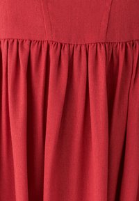 Vestito in tessuto rosso con superficie strutturata, caratterizzato da un corpetto aderente e una gonna ampia e drappeggiata con pieghe verticali.