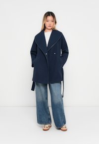Femme portant un manteau oversize bleu marine avec un large col, un jean large bleu clair et des baskets beiges, se tenant devant un fond blanc.