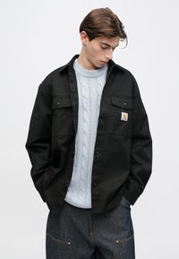 Czarna zapinana na guziki kurtka z dwoma kieszeniami na piersi, noszona na jasnoniebieskim swetrze z warkoczowym splotem, zestawiona z ciemnymi dżinsami.