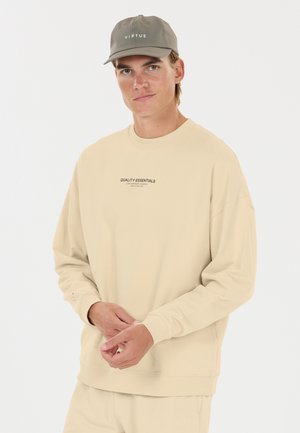 Jonge man draagt een beige sweatshirt met de tekst "Quality Essentials" en een grijze pet met het "Virtus"-logo, staand tegen een effen witte achtergrond.