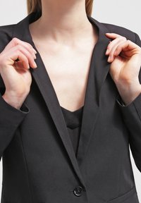 Žena upravuje klopy svého černého saku, což odhaluje výstřih černého vnitřního topu.