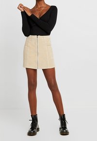 Gonna mini in corda beige con zip frontale e due tasche, indossata con una maglietta nera a maniche lunghe e stivaletti neri con lacci.