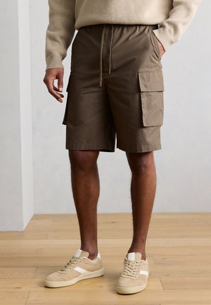 Mann trägt braune Cargo-Shorts mit Seitentaschen, beigefarbenen Pullover und beigefarbene Sneakers, steht auf Holzboden.