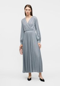 Γυναίκα που φοράει ασημί πλισέ μακρυμάνικο φόρεμα μέχρι τον αστράγαλο, κρατώντας μικρό ροζ τσαντάκι, φοράει μαύρες μυτερές γόβες, στέκεται πάνω σε λευκό φόντο.