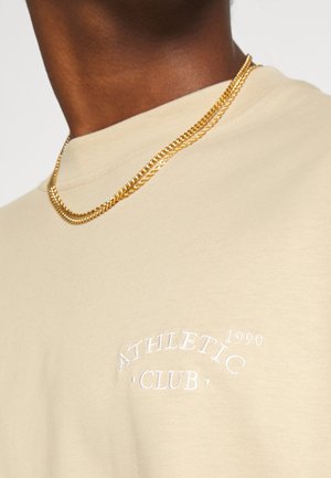 Gros plan sur une personne portant une chemise beige avec la broderie "ATHLETIC CLUB 1990" et deux colliers en chaîne dorée.