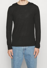 Mann in einem enganliegenden schwarzen Langarm-T-Shirt mit Rundhalsausschnitt und hellblauen Jeans, der vor einem schlichten weißen Hintergrund steht.