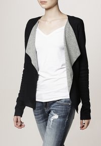 Svart cardigan med grå draperad front, långa ärmar och figurnära passform. Bärs över en vit V-ringad tröja och slitna blå jeans.