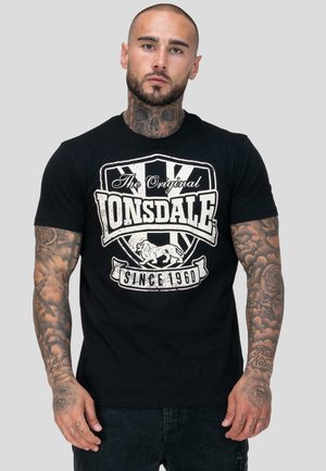 Hombre con tatuajes en el cuello y los brazos, vistiendo una camiseta negra de Lonsdale con logo y texto en blanco, de pie contra un fondo gris liso.