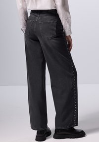 Jeans negros de pierna ancha y tiro alto con detalles de remaches a lo largo de las costuras laterales y dos bolsillos traseros. El material tiene una textura de denim suave.