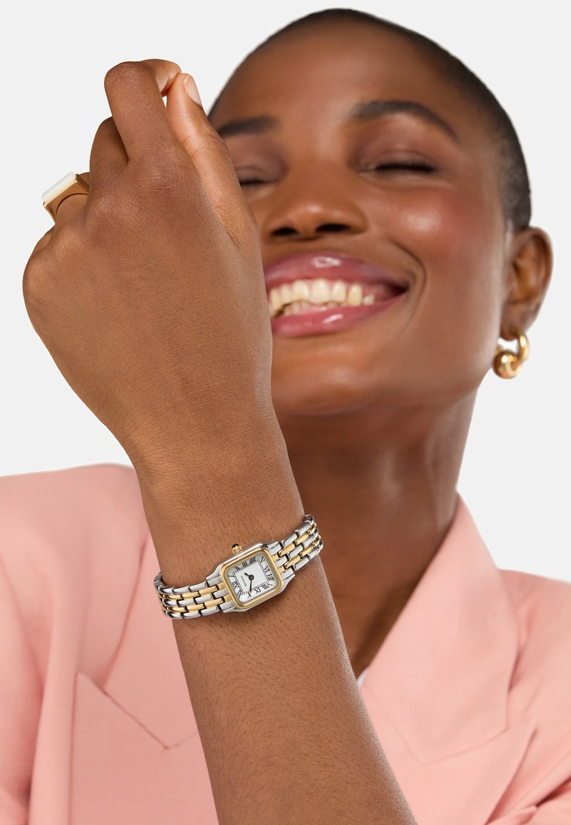 Montre-bracelet carrée avec un cadran blanc, un bracelet en métal doré et argenté, et des chiffres romains. Portée au poignet d'un modèle.