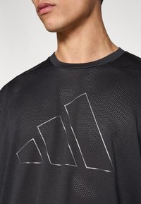 Μαύρο αθλητικό μπλουζάκι από δίχτυ με μεγάλο, λευκό περίγραμμα τριών στυλιζαρισμένων γεωμετρικών σχημάτων στο μπροστινό μέρος, στρογγυλή λαιμόκοψη και κοντά μανίκια.