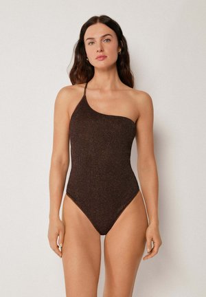 Vrouw met donker haar, die een éénschouder badpak draagt in donkerbruin met een getextureerd patroon, staand tegen een eenvoudige lichte achtergrond.