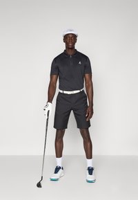 Svart kortermet polo-skjorte med glidelås, svarte shorts, hvit belte, hvit caps, og golf sko med blå detaljer; holder en golfklubbe.