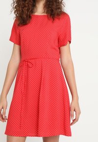Robe rouge à manches courtes avec des pois blancs, taille cintrée avec un lien, jupe évasée et tissu à la texture lisse.