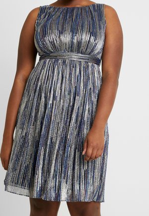 Mujer con un vestido plateado y azul sin mangas, plisado y hasta la rodilla, con una cintura ajustada sobre un fondo liso.