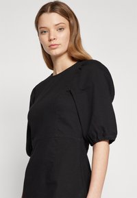 Robe noire avec de courtes manches bouffantes, un col rond et un corsage ajusté. Fabriquée en tissu lisse avec des détails de couture subtils.