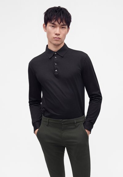 Homme portant un polo noir à manches longues boutonné et un pantalon vert foncé, debout les mains dans les poches devant un fond blanc.