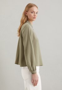 Olijfgroene blouse met lange mouwen en een relaxte pasvorm, knoopdetail bij de manchetten en een kraag. Gecombineerd met lichte broeken.
