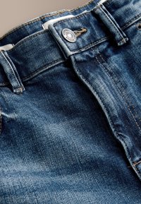 Zbliżenie na pas niebieskich dżinsów z denimu pokazujące metalowy guzik, szlufki na pasek i teksturowane przeszycia materiału.