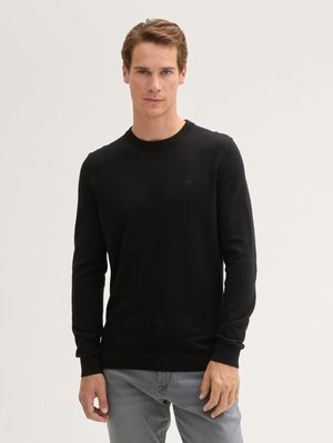 Man wearing black long-sleeve sweater and light gray jeans standing against plain light background.