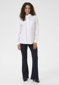 Camicia bianca con colletto rotondo, taschino sinistro, maniche lunghe; abbinata a jeans svasati scuri e scarpe nere a punta.