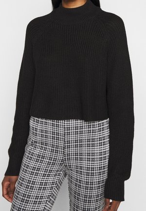 Personne portant un pull noir côtelé à manches longues et un pantalon à carreaux noir et blanc taille haute, debout devant un fond blanc.
