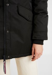 Parka noire isolée avec fermeture éclaire, poches à bouton-pression et poignets côtelés. Présente un cordon de serrage contrastant avec des rayures rouges et bleues.