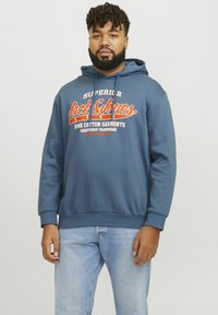 Blå bomulds hættetrøje med snøre i hætten, der har hvid og orange tekst på tværs af brystet. Sammen med lyseblå jeans.