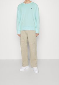 Sudadera verde menta con puños y dobladillo acanalados, combinada con pantalones beiges con manchas de pintura y un ajuste holgado. Zapatillas blancas completan el conjunto.
