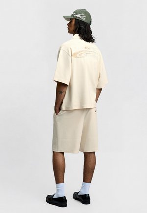 Homme aux cheveux mi-longs portant un ensemble beige texturé composé d'une chemise et d'un short, casquette verte, chaussettes blanches et chaussures noires, debout les mains dans les poches.