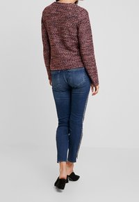 Pull texturé rouge et noir, design ajusté avec des manches longues. Jean skinny bleu avec des embellissements sur le côté. Mules noires à talons.