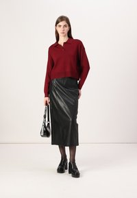 Czerwona dziergana koszulka polo z kołnierzykiem, połączona z czarną skórzaną spódnicą midi. Modelka trzyma czarną torbę i nosi masywne czarne buty na platformie.