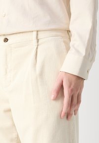 Beżowe spodnie sztruksowe z pionowymi żeberkami, z zapięciem na guzik i kieszeniami bocznymi. Na sobie ma jasną koszulę z długim rękawem, z podwiniętymi mankietami.