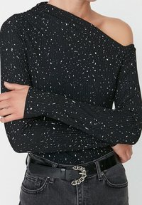 Svart ribbad långärmad topp med vita fläckar, en off-shoulder design och textilstruktur. Kombinerad med ett svart bälte och grå jeans.