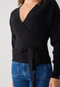Schwarzer gerippter Wickelsweater mit tiefem V-Ausschnitt und langen Ärmeln. Mit einem gebundenen Stoffgürtel in der Taille, kombiniert mit blauen Jeans.