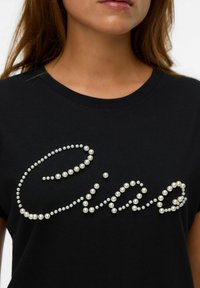 T-shirt noir à manches courtes arborant un design décoratif en perles blanches en forme de perles qui épellent "Ciao". Texture de tissu lisse.