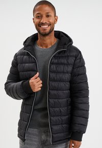 Chaqueta acolchada negra con capucha desmontable, cierre de cremallera y puños de canalé. Presenta costuras verticales y una textura suave y brillante.
