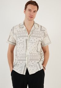 Camisa de manga corta en tela beige claro con patrones geométricos negros y motivos sutiles, con un cierre de botones en la parte delantera y cuello.