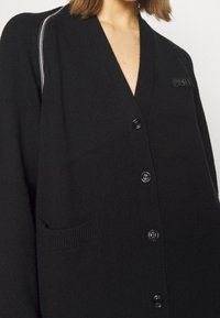 Svart stickad cardigan med v-ringning, framknappar, stora fickor och en märkespatch. Har en texturerad yta och avslappnad passform.