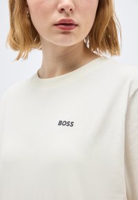 Νεαρό άτομο με κοντά, ανοιχτό καστανά μαλλιά, που φοράει λευκό μπλουζάκι με στρογγυλή λαιμόκοψη και τη λέξη "BOSS" τυπωμένη με μικρά μαύρα γράμματα στο στήθος.