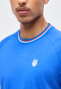 Νέος άνδρας που φοράει μπλε πουκάμισο K-Swiss με κόκκινο, λευκό και μπλε ριγωτό γιακά, εμφανιζόμενος από τον λαιμό έως το πάνω μέρος του στήθους.