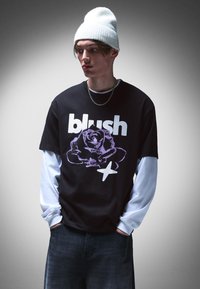 Black t-shirt with white text "blush" and purple rose graphic. Layered with a light blue long-sleeve shirt underneath. Textured white beanie.