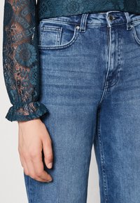 Džínové kalhoty ve středně modré barvě s přiléhavým střihem. Lehce strukturovaná látka s viditelným štepováním a kovovými knoflíky v oblasti pasu.