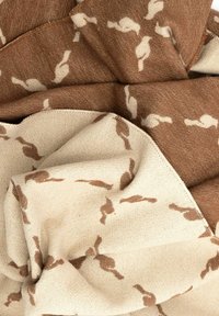 Brązowa i beżowa dzianina z powtarzającym się wzorem małych sylwetek ptaków w kontrastujących kolorach, pokazująca złożone i nałożone warstwy.