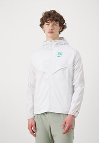 Mladý muž s kučeravými vlasmi, oblečený v bielom zipsovom mikinovom kabáte s kapucňou a svetlozelených nohaviciach, stojí na pozadí v jednoduchom bielom prostredí.