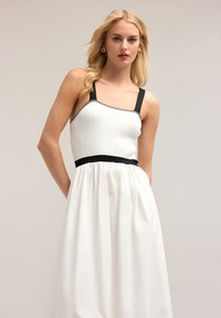 Rochie albă cu un corset ajustat și nervurat și bretele negre, având o talie contrastantă de culoare neagră. Fustă dintr-un material neted și curgător.