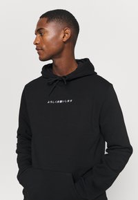 Black hoodie with a drawstring, featuring white text in Japanese on the chest. Made of a soft, textured fabric. Adjustable cuffs.