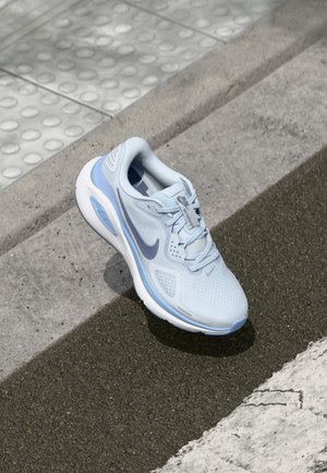 Lichtblauwe Nike hardloopschoen met witte zool, rustend op asfalt vlakbij detecteerbare tegel en stoeprand.